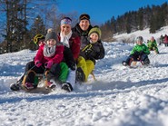 Eine Familie mit drei Kindern fährt mit einem Schlitten den Hügel herunter, dahinter weitere Kinder auf Schlitten.