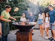 Eine Frau steht am Grill und brät Burger, Kinder und Erwachsene schauen zu.
