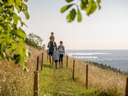 Eine Familie spaziert auf einem Pfad zwischen Wiesen, im Hintergrund ein See.