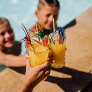 Zwei Kinder im Pool und eine weitere Person stoßen mit fruchtigen Getränken an.