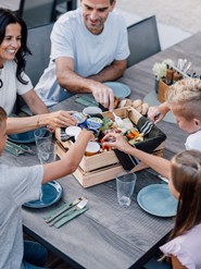 Familie sitzt an einem Tisch & langt in eine Kiste gefüllt mit regionalen Spezialitäten.