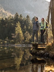 Zwei Wanderer stehen am Ufer eines Sees der Umgeben ist von Bäumen und Bergen.