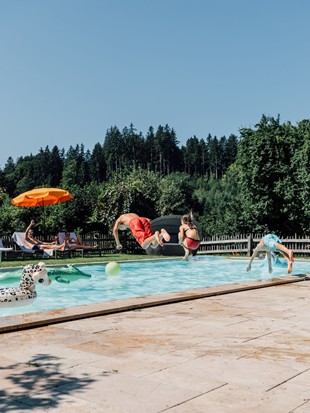 Kinder springen in den Pool, die Eltern liegen auf Liegestühlen und schauen zu.