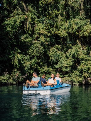 Eine Familie auf einem Tretboot am See, am Ufer stehen hohe Nadelbäume.