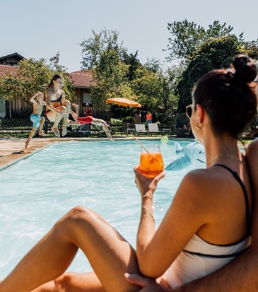 Ein Mann & eine Frau sitzen Am Beckenrand mit Getränk in der Hand, Kinder springen in den Pool.