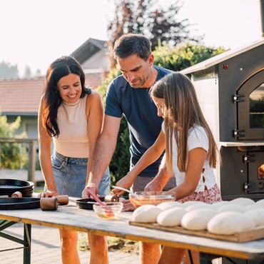 Eine Familie vor einem großen Tisch mit Teig und Rollern, das Kind rollt Pizzateig aus.