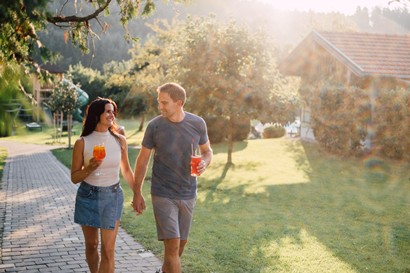 Ein Paar geht Hand in Hand mit Getränken in der Hand einen Pfad am Huberhof entlang.
