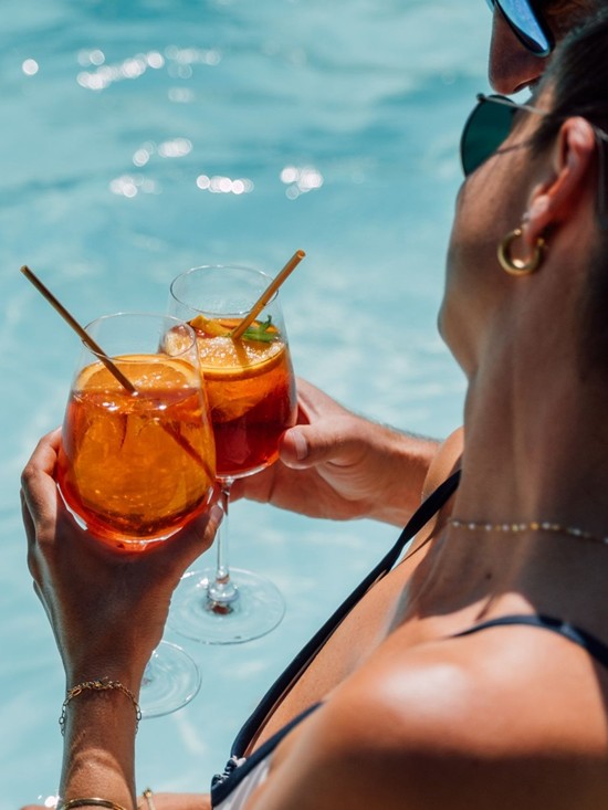 Ein Mann und eine Frau stoßen am Beckenrand des Pools mit Aperol an.