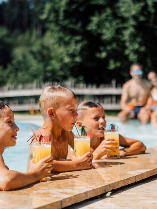 Drei Kinder im Pool trinken ein fruchtiges Getränk am Beckenrand, die Eltern sitzen im Hintergrund.