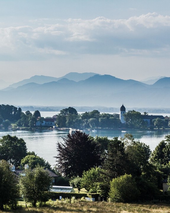 Ein See mit vielen Bäumen und Häusern am Ufer, weiter hinten sind Berge.